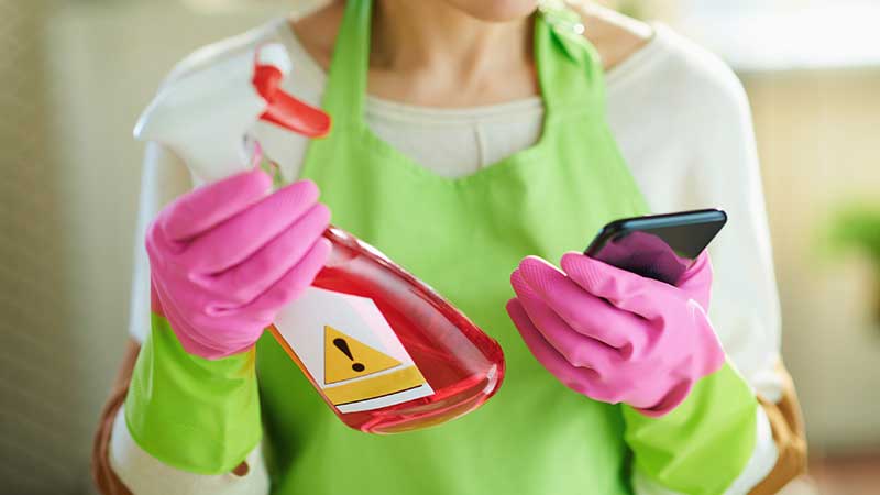 Mulher vestida com camisola branca e avental verde-alface, de luvas cor de rosa, a segurar a embalagem de um detergente vermelho com vaporizador numa mão e o telemóvel na outra